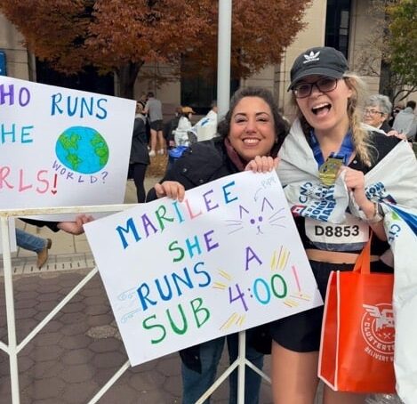 Two people smile and hold handmade marathon signs while standing behind a metal barrier. One person is wrapped in a finisher’s blanket with a race bib and medal.